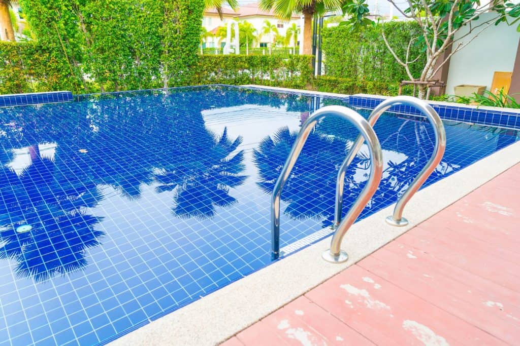 Beautiful backyard swimming pool with vibrant blue tiles, stainless steel ladder, surrounded by lush green hedges, palm trees, and a cozy outdoor patio for relaxation and entertainment.
