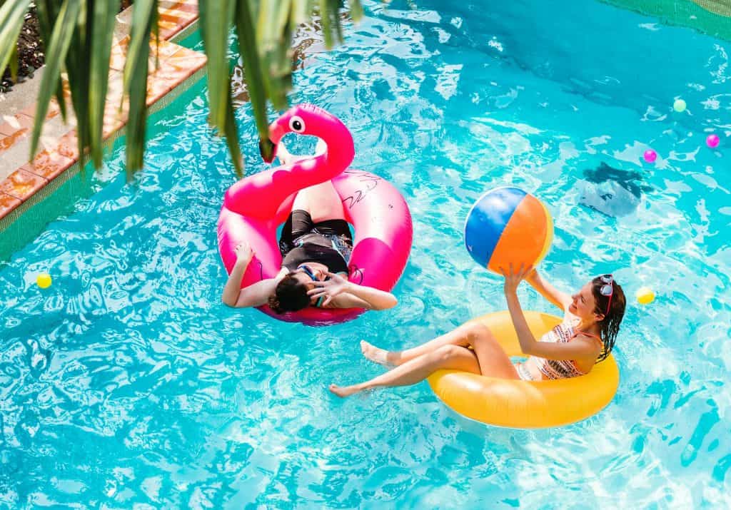Relaxing pool scene with two women enjoying summer on inflatable floats, one with a flamingo float and the other with a yellow ring, playing with a beach ball in a vibrant backyard pool.