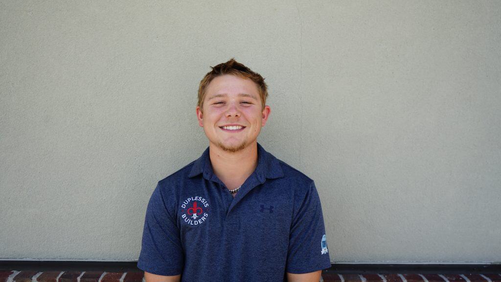 A friendly young man in a blue polo shirt with Duplessis Builders logo, smiling confidently against a plain wall.
