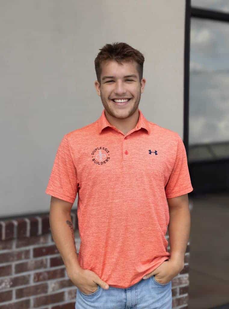A smiling young man in a coral polo shirt with the Duplessis Builders logo, standing outdoors in front of a modern building.