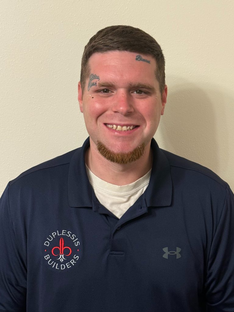 A professional pool builder from Duplessis Builders, smiling in a navy polo shirt with company logo, standing against a plain wall.