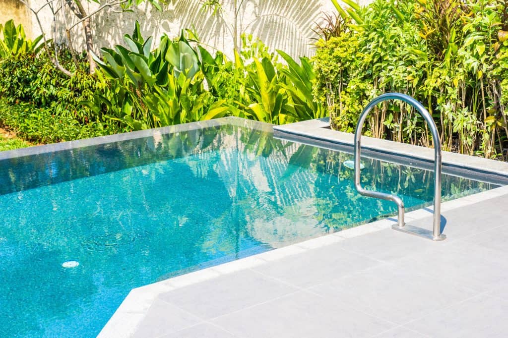 An inviting backyard pool featuring clear blue water, surrounded by a sleek white deck and vibrant tropical plants, perfect for relaxing and outdoor entertaining.