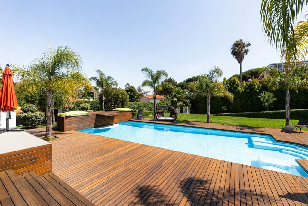Spacious outdoor pool area featuring a wooden deck, tropical plants, and shaded seating, perfect for relaxing and entertaining in a private backyard setting.