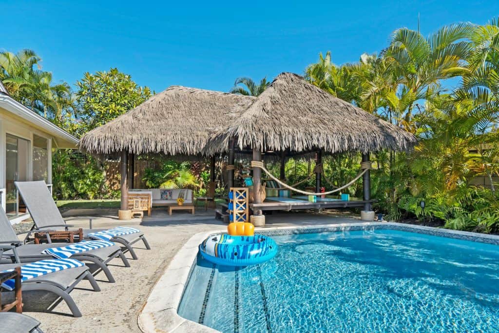 An inviting backyard pool featuring a thatched tiki hut, lounge chairs, and lush tropical landscaping, showcasing the expertise of a luxury pool builder in creating custom outdoor retreats.
